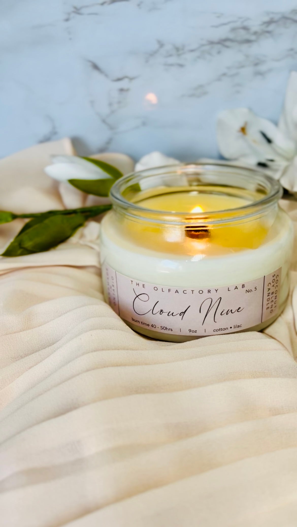 A lit Cloud Nine candle on a beige material against a marble background.