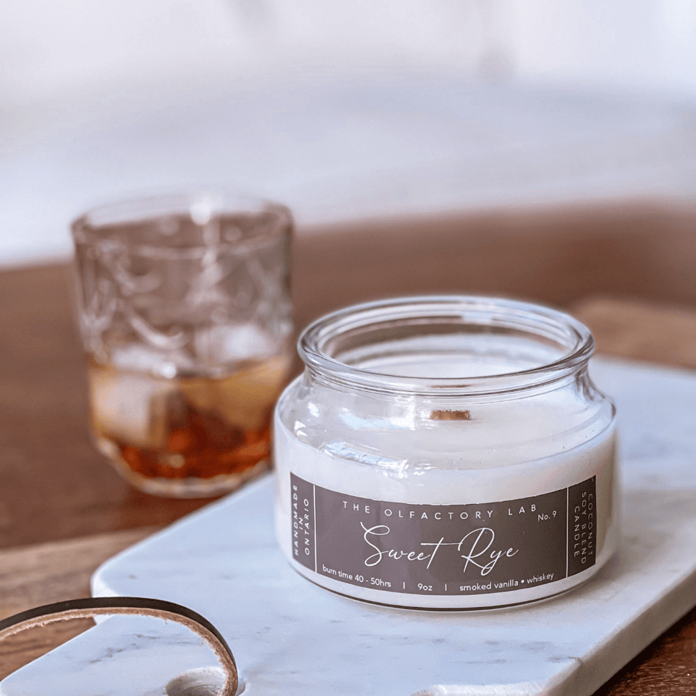 9oz white candle. Candle label is a rectangle shape in a dark brown colour with white cursive font reading Sweet Rye. Candle is placed on a white marble cutting board with a glass of whiskey in the background.