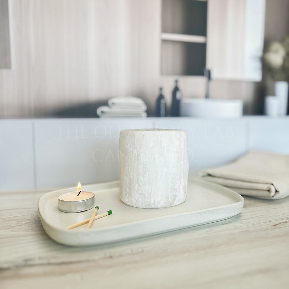
                  
                    Selenite holder on a white tray with a lit tea light and set of matches beside it to demonstrate the tea lights purpose. In the background is a luxury bathroom scape and a folded beige towel. 
                  
                