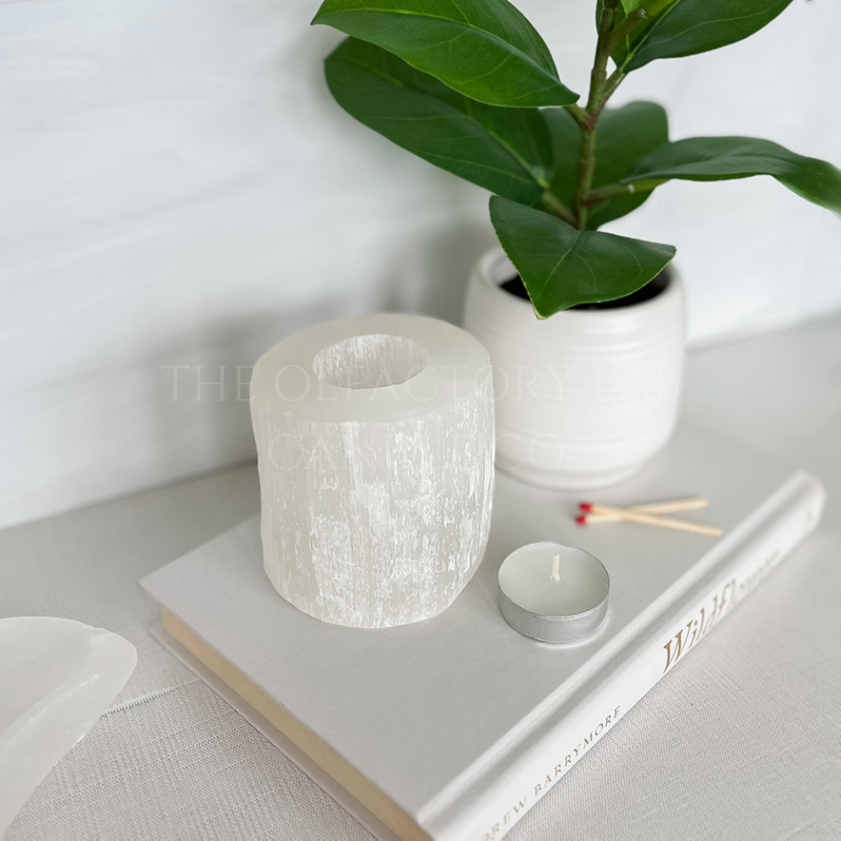 Selenite Tea Light Holder on a white hardcover book beside a green plant. In front of the holder is a tea light and matches to demonstrate the purpose of the tea light holder. 