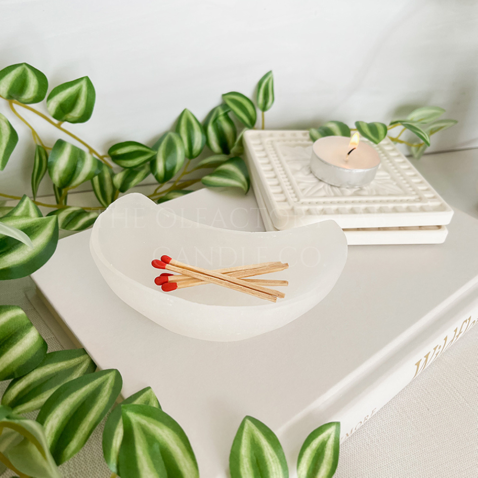 
                  
                    Selenite moon bowl pictured sitting on top of a book near some vines. Small tea light burning in the back corner
                  
                