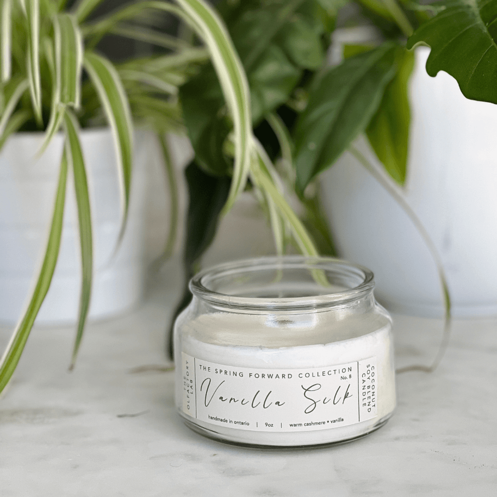 9oz white candle. The candle label is a rectangle shape in a soft yellow shade with grey cursive font reading Vanilla Silk. The candle is placed on a white marble counter with greenery in the background.