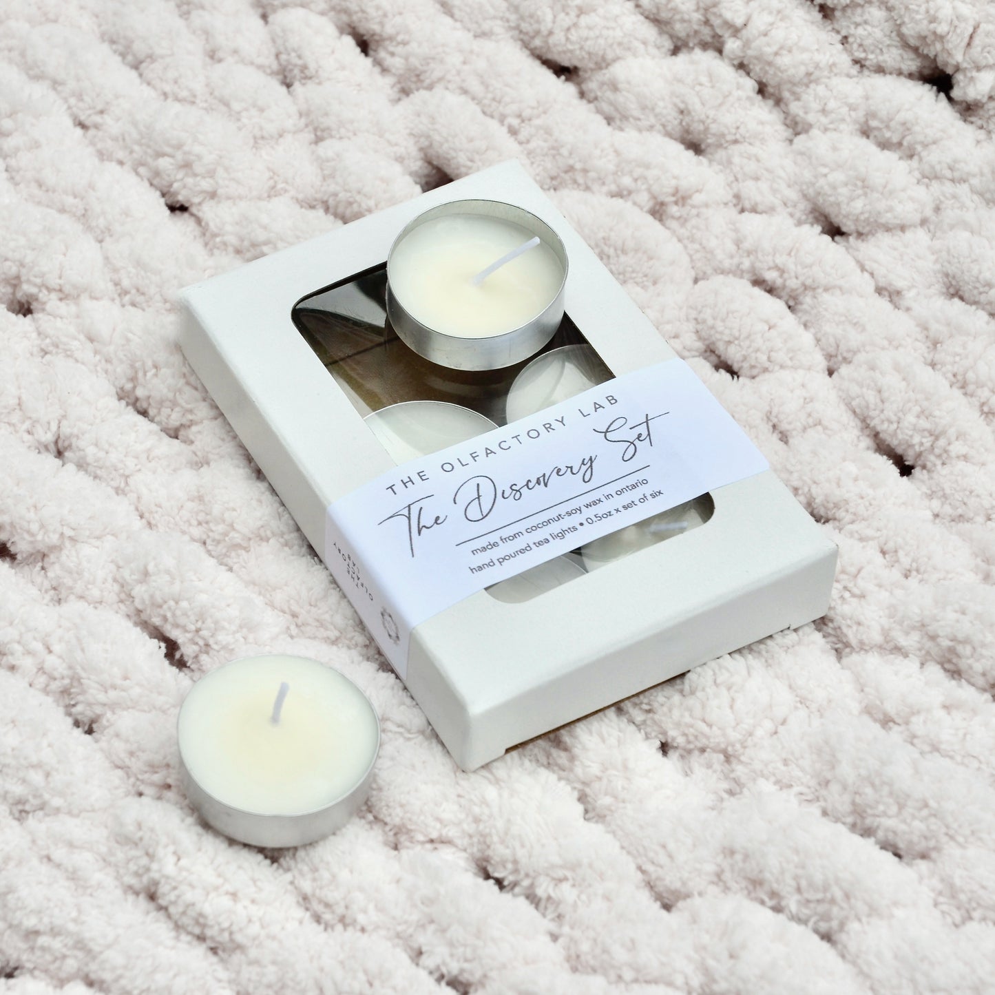 A white box with 6 tea lights. The box has a white rectangle label with black cursive font that reads The Discovery Set. The Discovery Set is placed on a white knit blanket with 2 tea lights outside of the box to display the tea lights.