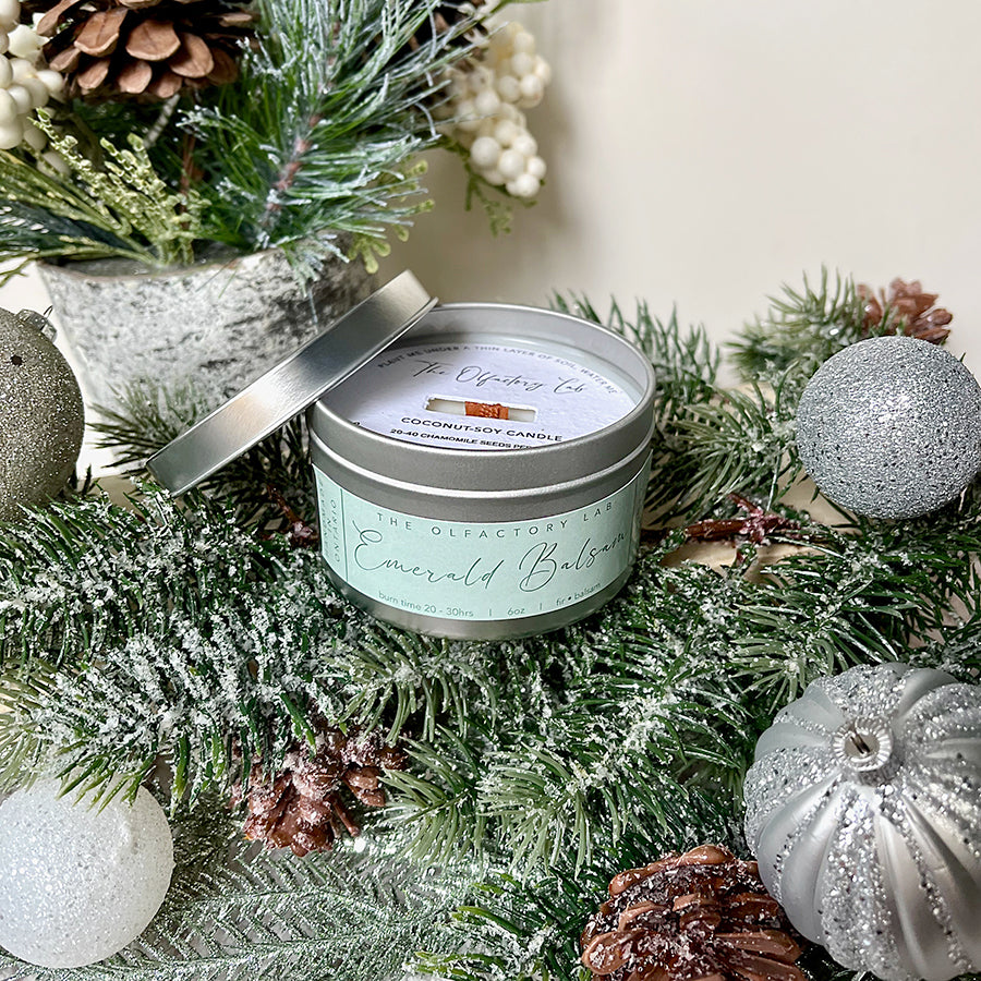
                  
                    6oz white candle in a silver tin. The candle has a square soft green label reading Emerald Balsam. The candle is sitting on a bed of pine greenery with silver ornaments surrounding it.
                  
                