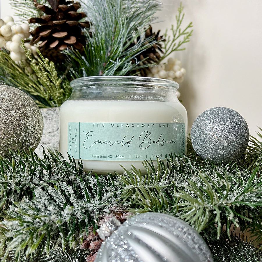 
                  
                    9oz white candle with a rectangle soft green label reading Emerald Balsam. The candle is sitting on pine greenery with silver ornaments around the candle.
                  
                