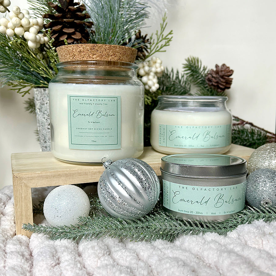 
                  
                    A trio of the 15oz, 9oz and 6oz  Emerald Balsam candles. The candles are sitting on a bed of pine greenery with silver ornaments surrounding the candles.
                  
                