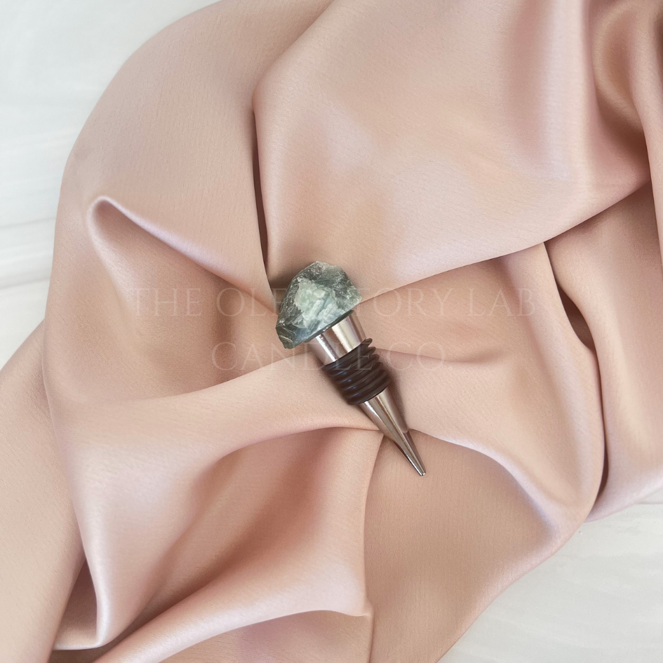 
                  
                    Silver wine stopper with a green Fluorite crystal top. The wine stopper is displayed in the middle of a soft pink silk background.
                  
                