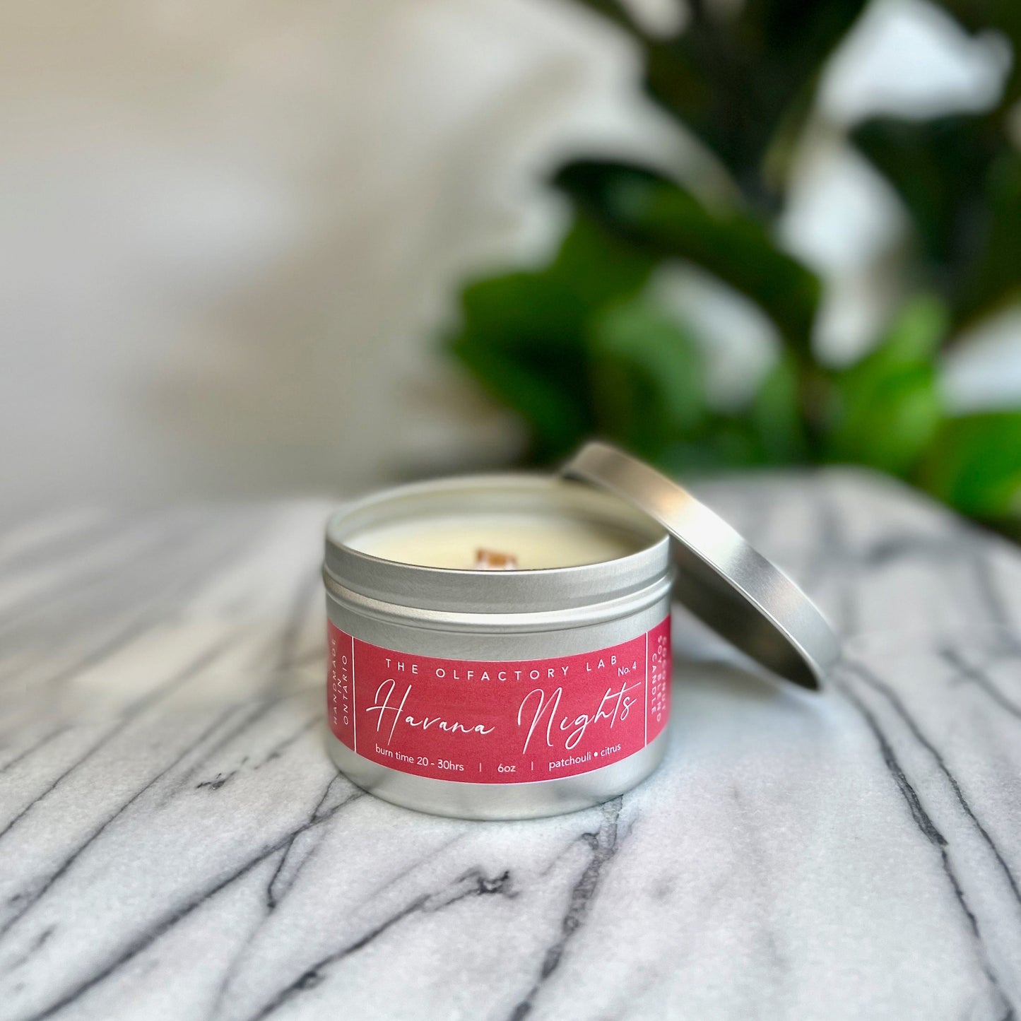 
                  
                    6oz Havana Nights candle in a silver tin. Label is a rectangle shape in a true red colour with white  cursive font. The candle is placed in the middle of a grey marble table with greenery in the background. Lid is placed off the right hand side of the handle to show the wood wick. 
                  
                