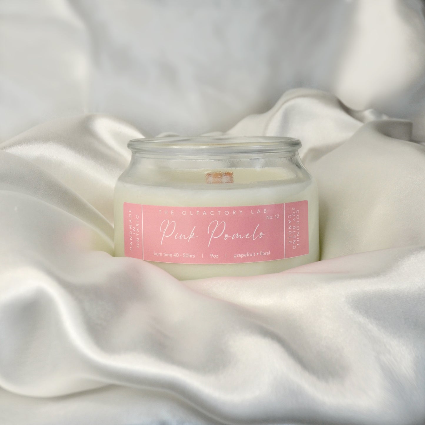 9oz white candle. Candle is laying on a white silk pillow. Candle label is a rectangle shape in a soft pink with white cursive font reading Pink Pomelo.