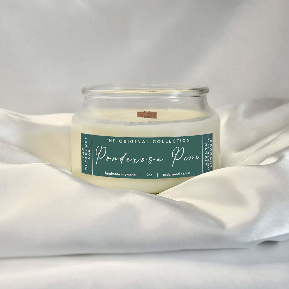 9oz white candle. Candle has a rectangle shaped label in a forest green with white cursive font reading Ponderosa Pine. The candle is sitting on a white silk pillowcase.