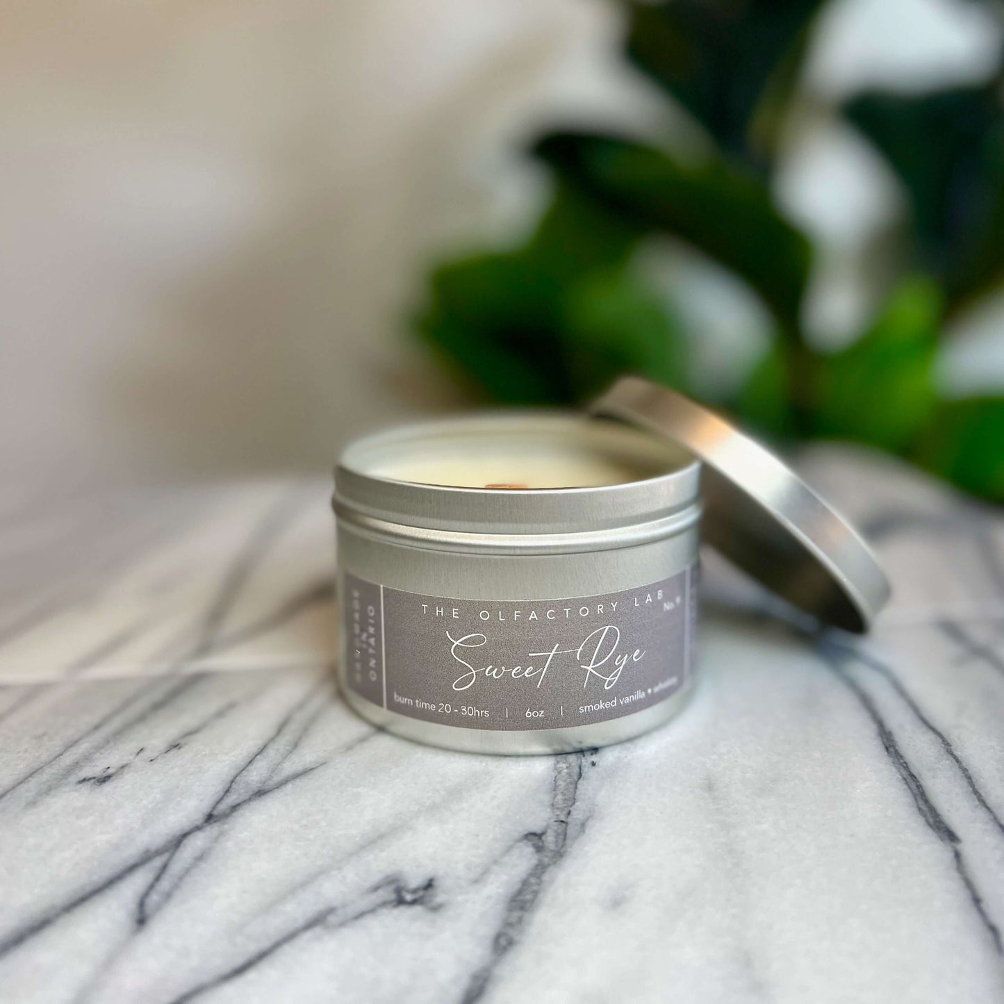 
                  
                    6oz candle in silver tin. Candle has a rectangle shaped label in a soft grey colour with white cursive font reading Sweet Rye. The candle is sitting on a grey and white marble table with greenery in the background. The candle lid is placed on the right side of the candle to display the wood wick.
                  
                