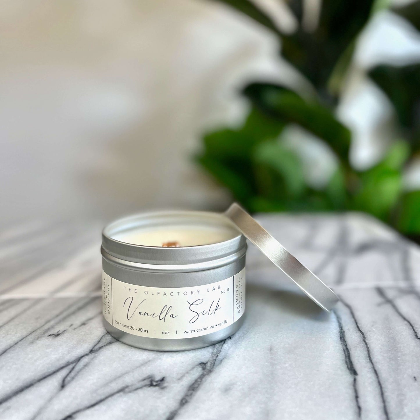 
                  
                    A 6oz candle in a silver tin. The candle label is a rectangle shape in a soft yellow with grey cursive font reading Vanilla Silk. The candle is displayed on a white and grey marble table with greenery in the background. The candle lid is placed on the right side of the candle to display the wood wick.
                  
                
