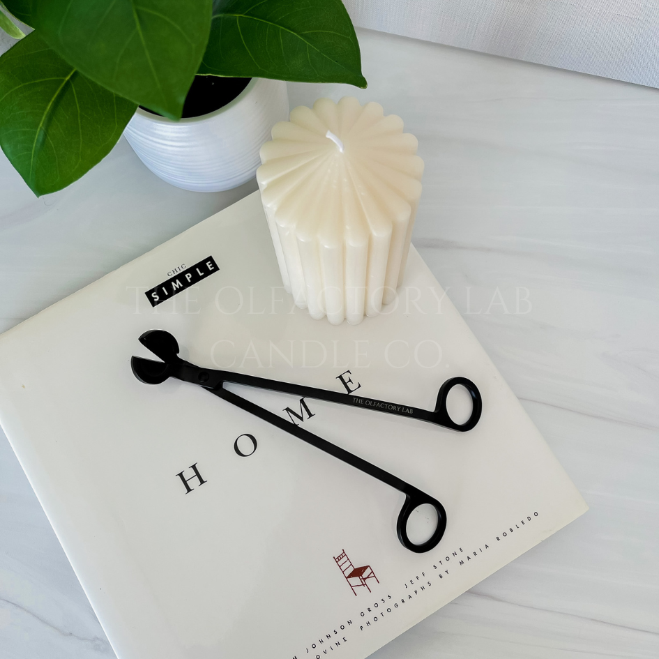 A black wick trimmer on a white hardcover book that reads HOME. The wick trimmer is beside a decorative pillar candle and greenery.