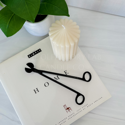 A black wick trimmer on a white hardcover book that reads HOME. The wick trimmer is beside a decorative pillar candle and greenery.
