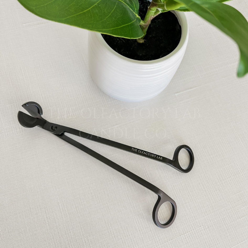 
                  
                    A black wick trimmer on a beige surface in front of a green decorative plant. Engraved in the wick trimmer is The Olfactory Lab. 
                  
                