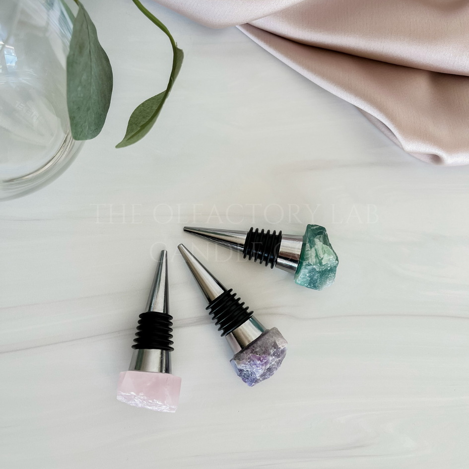 3 crystal wine stoppers on a grey marble surface. The wine stoppers are silver with a crystal top in Rose Quartz (soft pink), Amethyst (purple), and Fluorite (green/purple).  